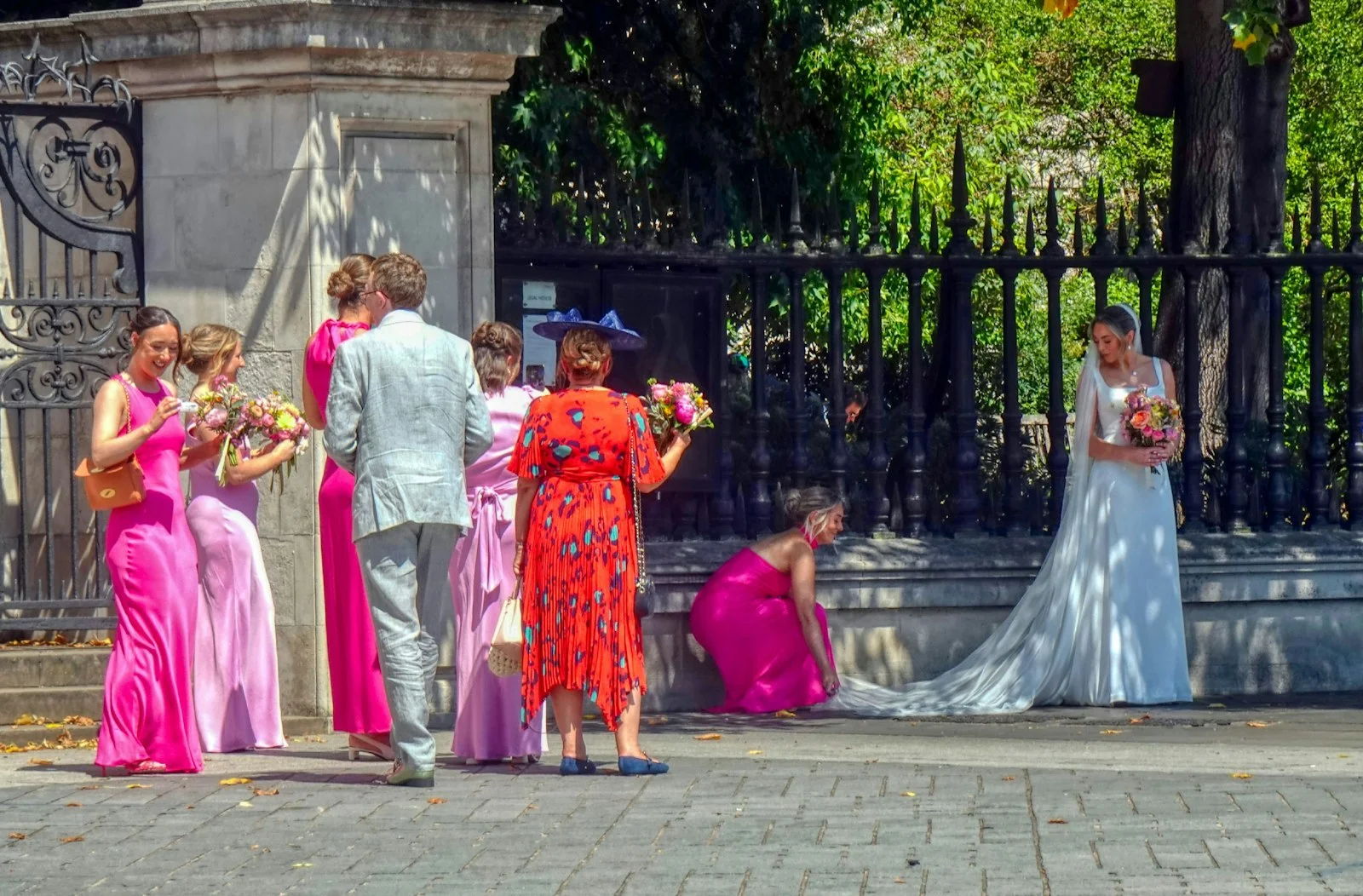 A bride in a white gown and guests in colorful dresses near a metal fence.